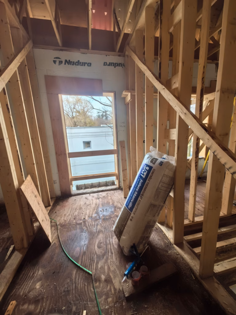 A construction site features wooden beams framing walls and a large, open doorway, hinting at the luxury homes custom builders are crafting. Insulation materials and a black hose lie on the unfinished floor. Through the doorway, a tree and part of another building in Toronto are visible.
