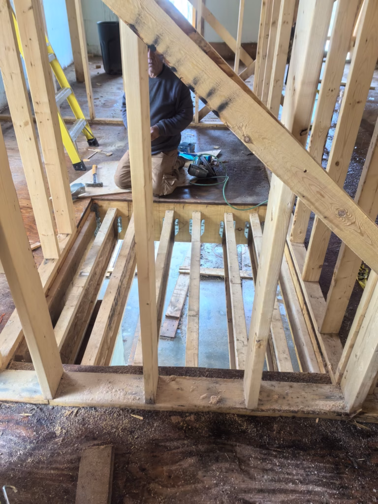 A person is kneeling on the floor, working on a wooden structure with vertical beams and a partially constructed floor. Tools are scattered about, and a yellow ladder stands in the background. The setting suggests custom built homes Toronto, embodying precision and craftsmanship at its core.