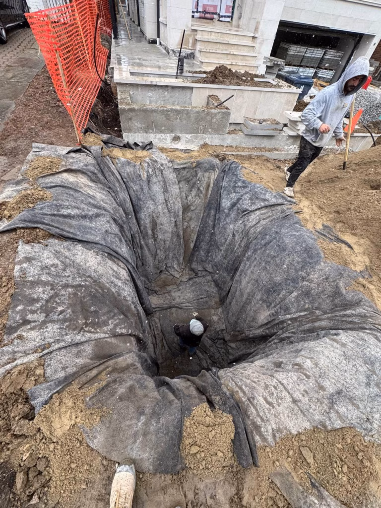 A deep, circular construction hole lined with fabric, with a worker inside and another standing nearby, a red safety fence in the background.