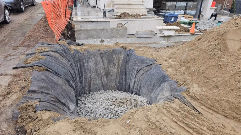 A deep, gravel-filled excavation surrounded by black landscaping fabric, with construction materials and workers in the background.