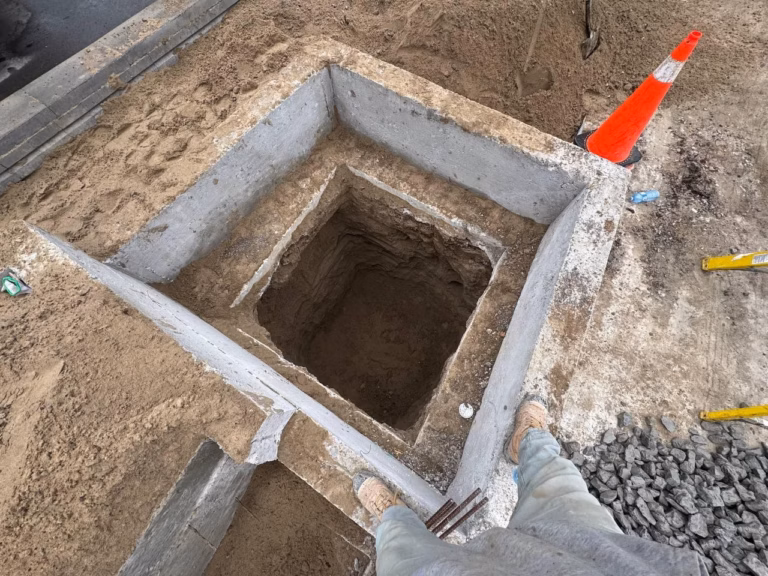 A construction site showing a square concrete foundation hole surrounded by sand, gravel, and a traffic cone. Workers' feet are visible.