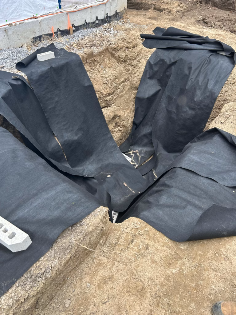 A deep construction pit with black landscape fabric laid in folds, surrounded by gravel, and a concrete block resting on the edge.