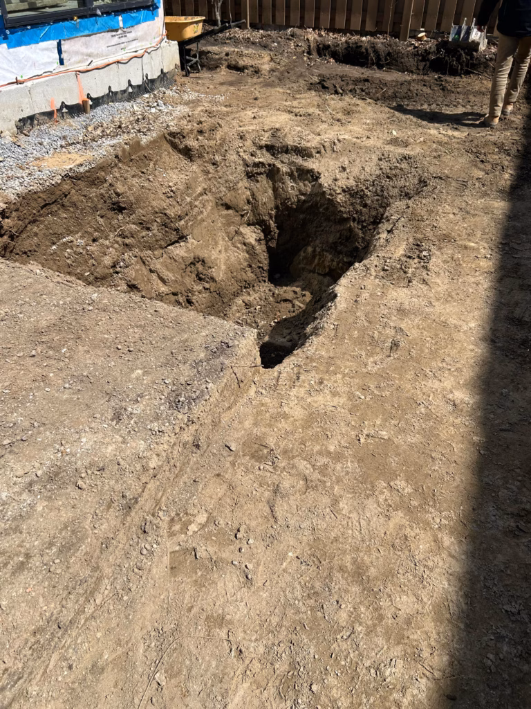 \A large, deep excavation in brown dirt, with a wheelbarrow nearby, showing preparations for construction near a building foundation