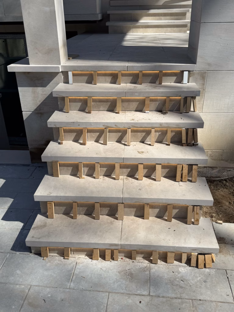 A set of unfinished stone stairs with wooden supports underneath each step, positioned outside a modern building.