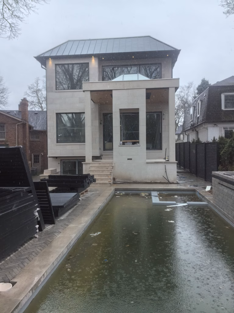 A modern two-story house with a metal roof, featuring large windows, in a snowy environment, alongside a pool with murky water.