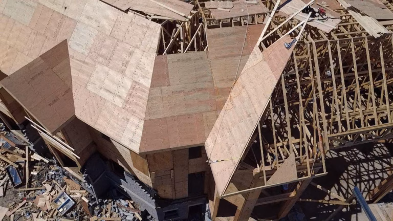 Aerial view of a partially constructed building featuring wooden framing and plywood roof panels, surrounded by construction debris.