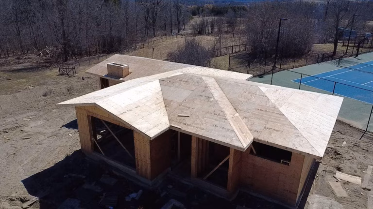 Aerial view of a partially constructed house with a multi-angled roof, surrounded by bare trees and a nearby tennis court.