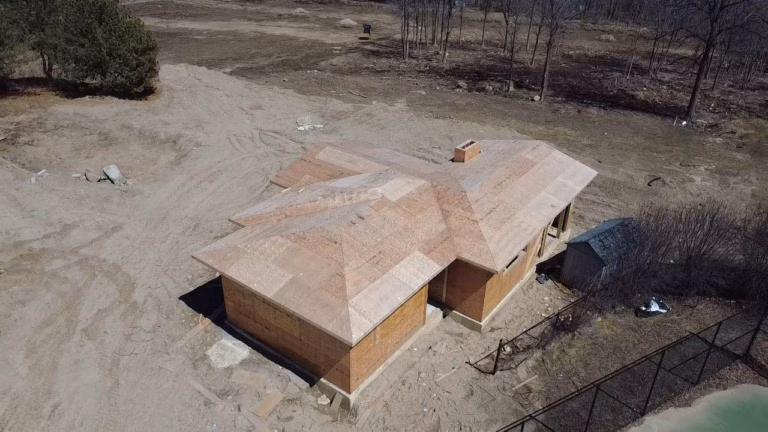Aerial view of a partially constructed house with a multi-angled roof, surrounded by bare trees and a nearby tennis court.