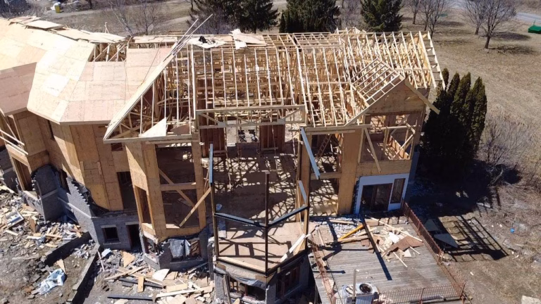 An aerial view of a partially constructed house with exposed wooden framing, debris scattered around, and a clear sky in the background.