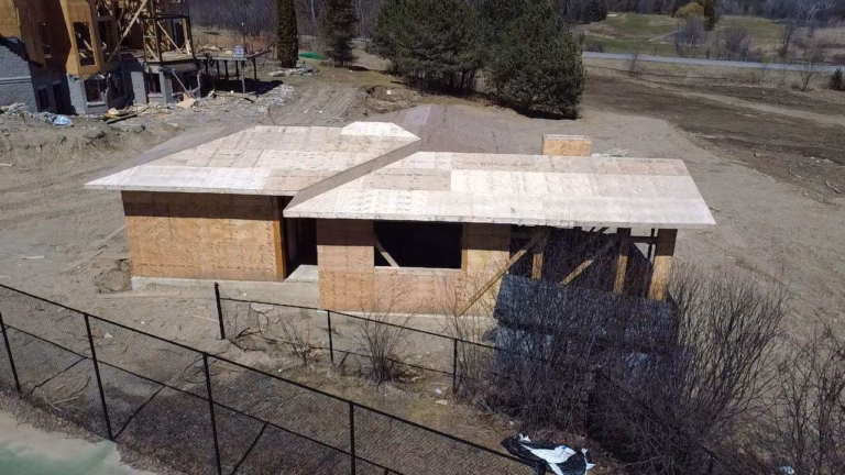 Aerial view of an unfinished wooden house with a plywood roof, surrounded by dirt, bushes, and partially constructed neighboring buildings.
