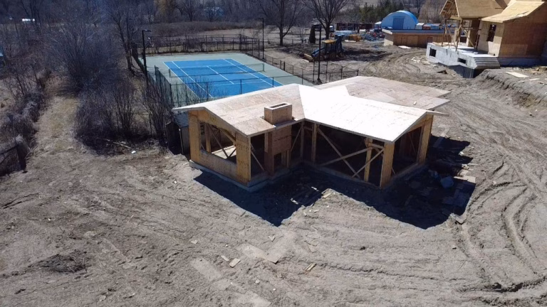 Aerial view of an under-construction house beside a blue tennis court, surrounded by bare trees and a sandy landscape.