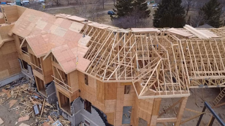 Aerial view of a partially constructed house featuring wooden framing and plywood sheathing on the roof, surrounded by construction debris.