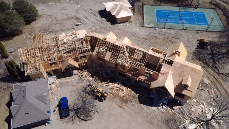 Aerial view of a partially constructed house with exposed wooden framing, surrounded by construction debris and a tennis court nearby.