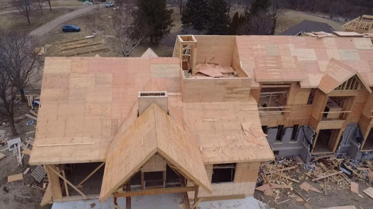 Aerial view of a large residential building under construction, featuring wooden frameworks, a partially completed roof, and surrounding debris.