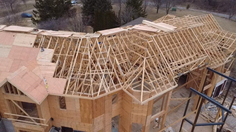 Aerial view of a wooden house under construction, showcasing the framing and roof structure with surrounding trees and vehicles.