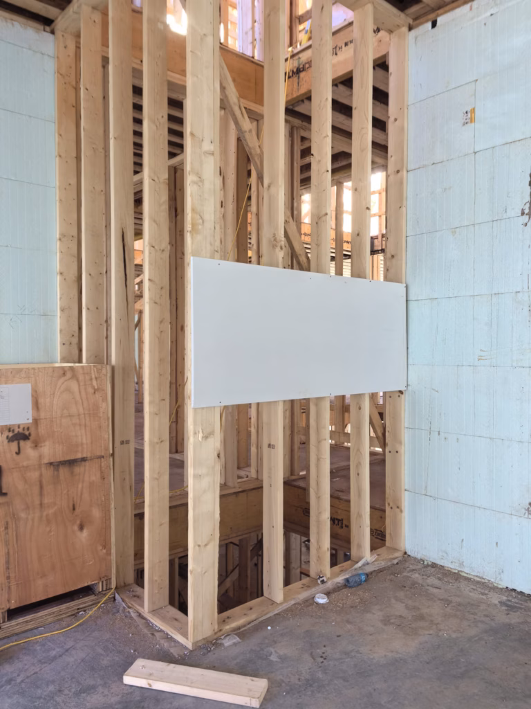 A partially constructed wall with exposed wooden framing and insulation panels in a building under construction, typical of custom-built homes in Toronto. A white rectangular board is attached to the wood structure, while the concrete floor is dotted with scattered construction materials.